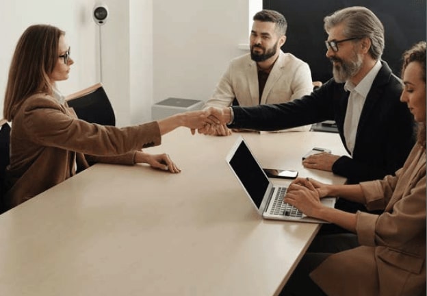 Four people sitting at meeting table - two people are shaking hands.