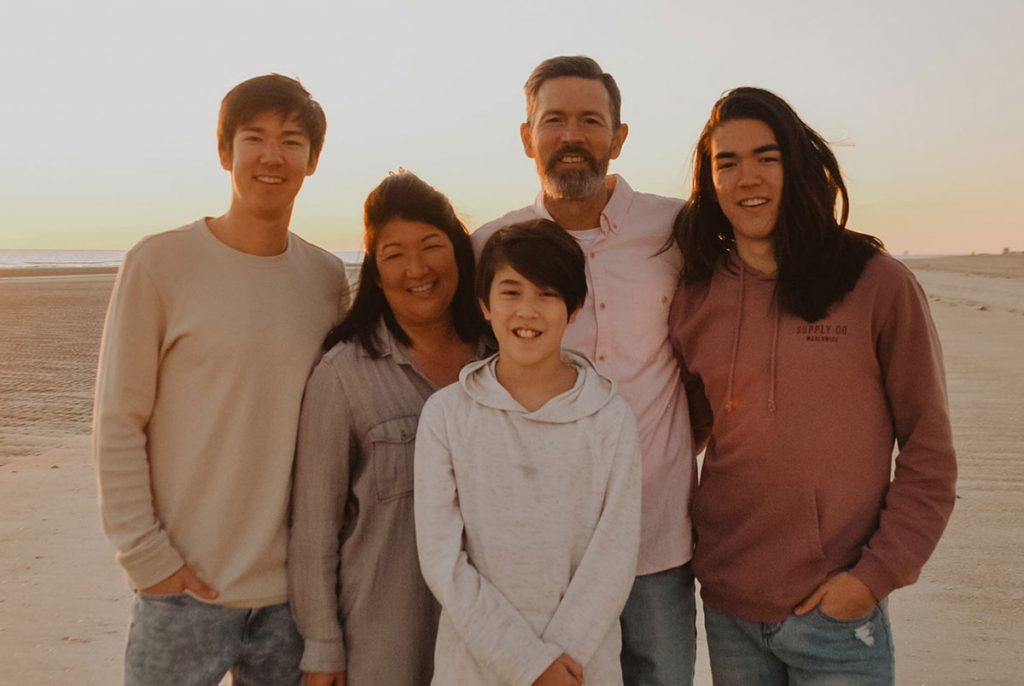 Parents and 3 boys standing on sand and sunset all facing the camera