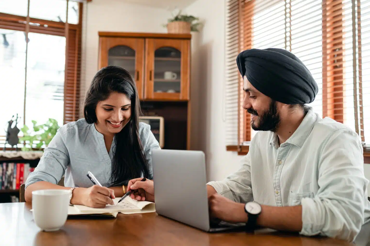 Woman and man sitting at dining room table, man in front of computer and woman writing in a notebook