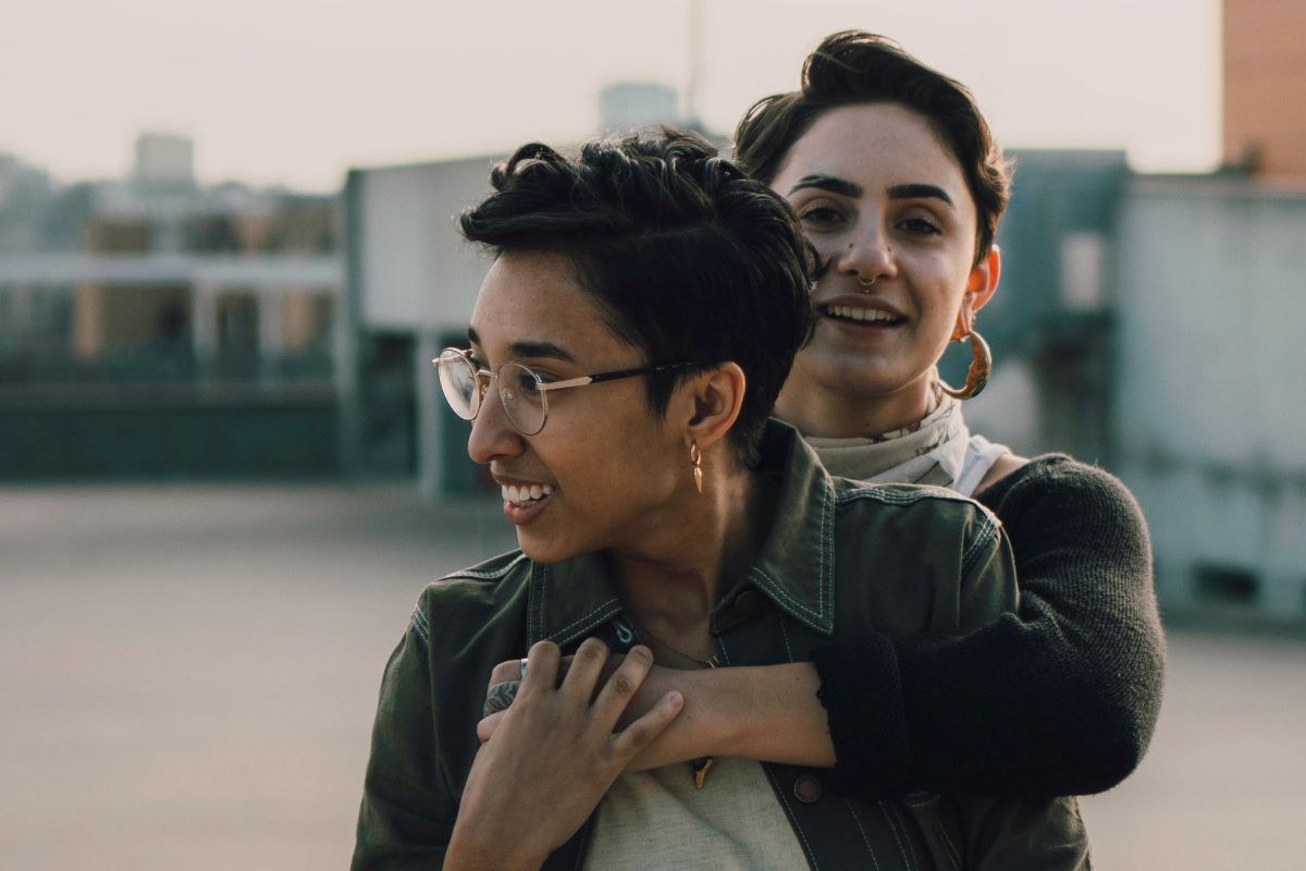 Couple with one standing behind with her arms around her girlfriend who might celebrate Valentine's Day.