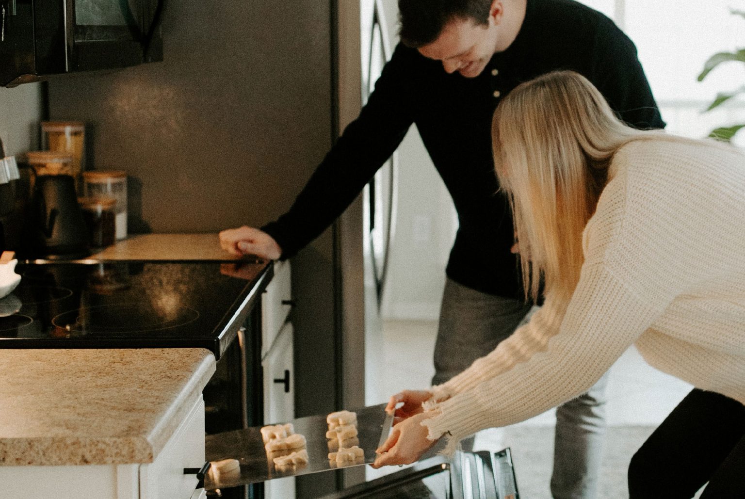 Woman leaning down to put a tray in the over. Man leaning on counter top also looking into oven