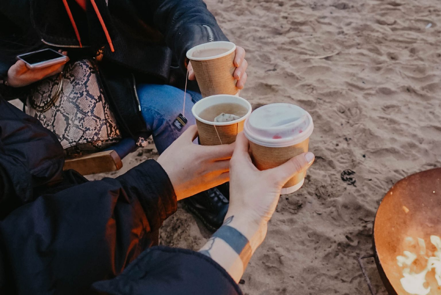 hands holding coffee cups near a fire pit