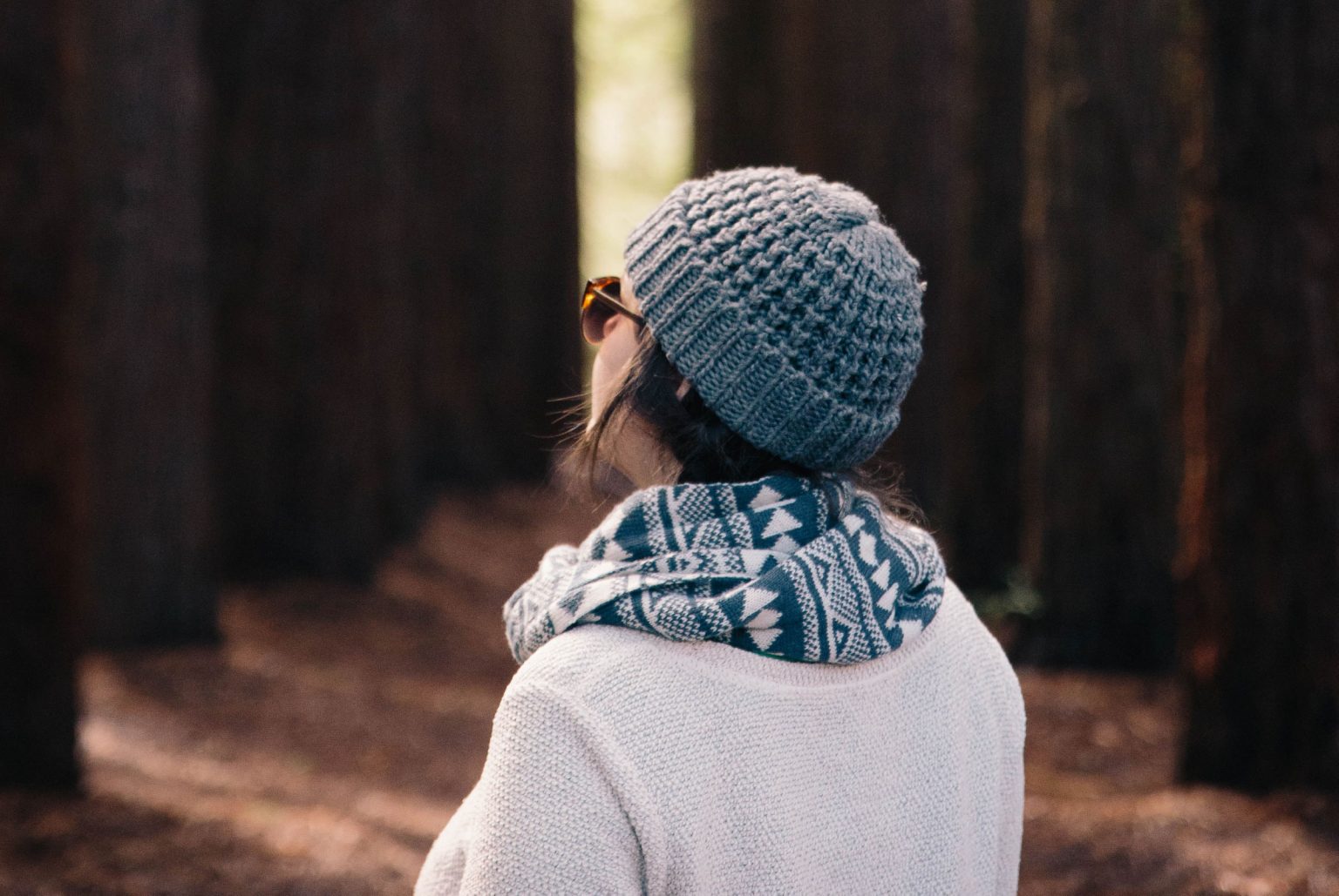 Woman wearing beanie, scarf and suglasses facing away from the camera in a forest setting