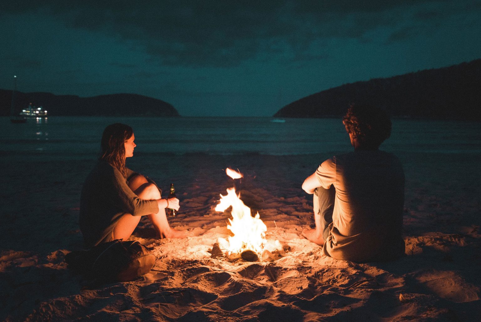 Two people sitting next to a campfire on the beach at dusk