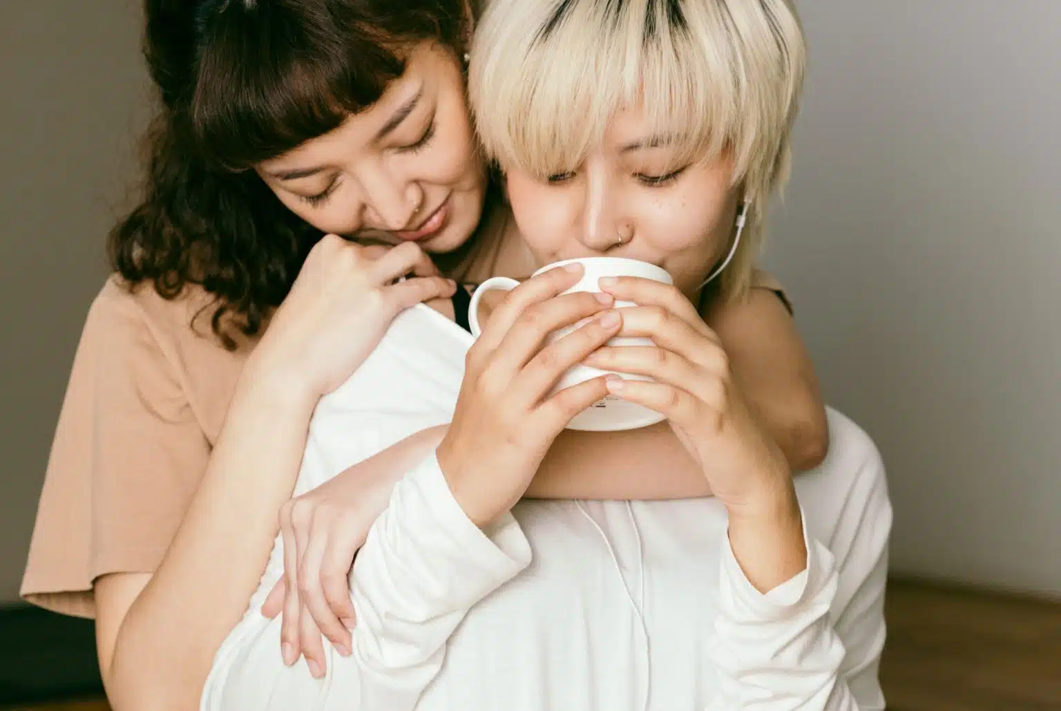 Woaman hugging another woman from behind. One sips from a mug
