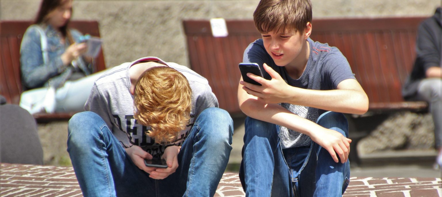 Two boys on smartphones, one crouched with head between legs, highlighting social media safety focus.