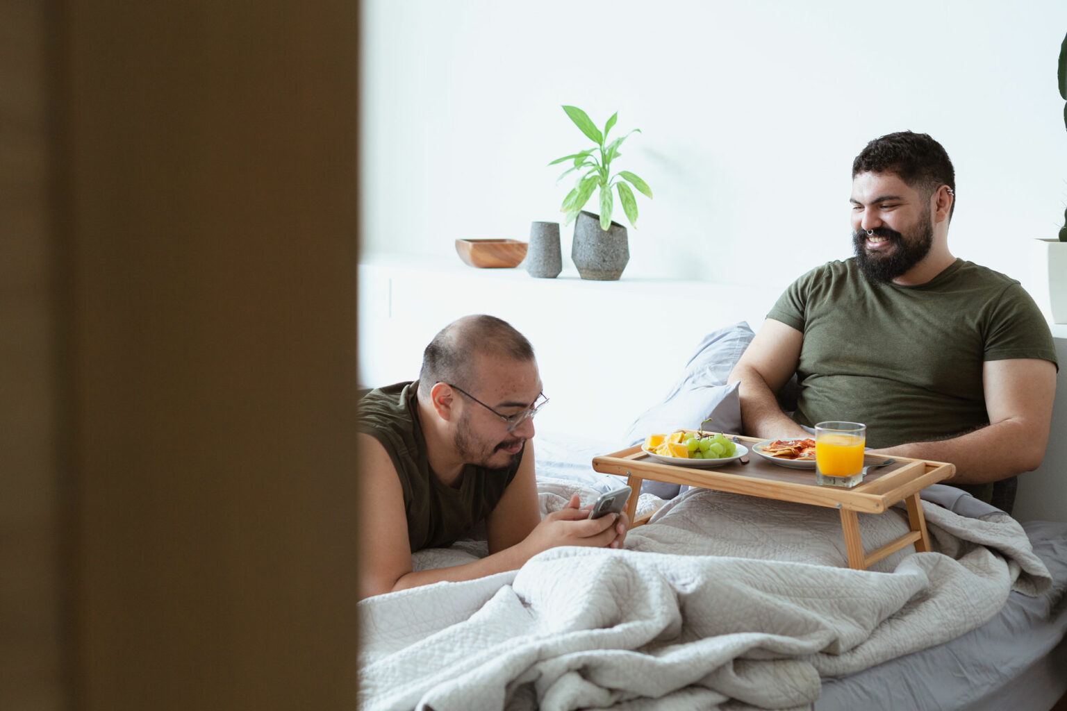 Happy LGBTQIA+ couple enjoying breakfast in bed together, one checking his phone while the other eats breakfast, showcasing a loving relationship free from LGBTQIA+ discrimination