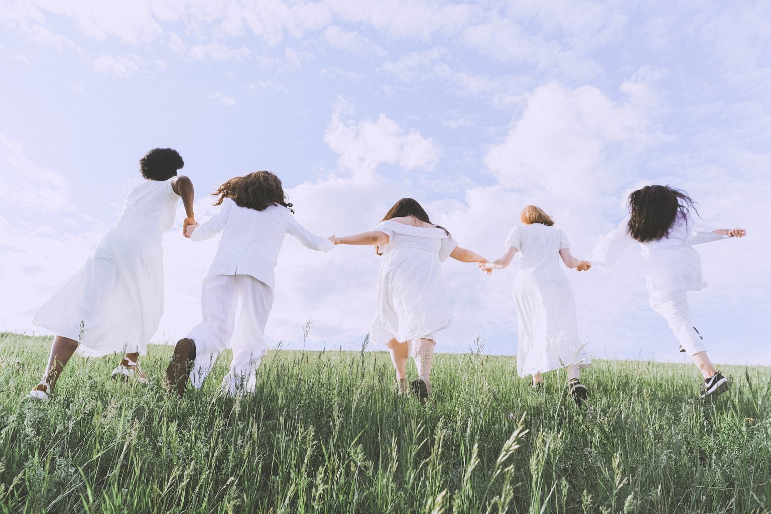 Five women running across a field of grass all wearing white and holding hands