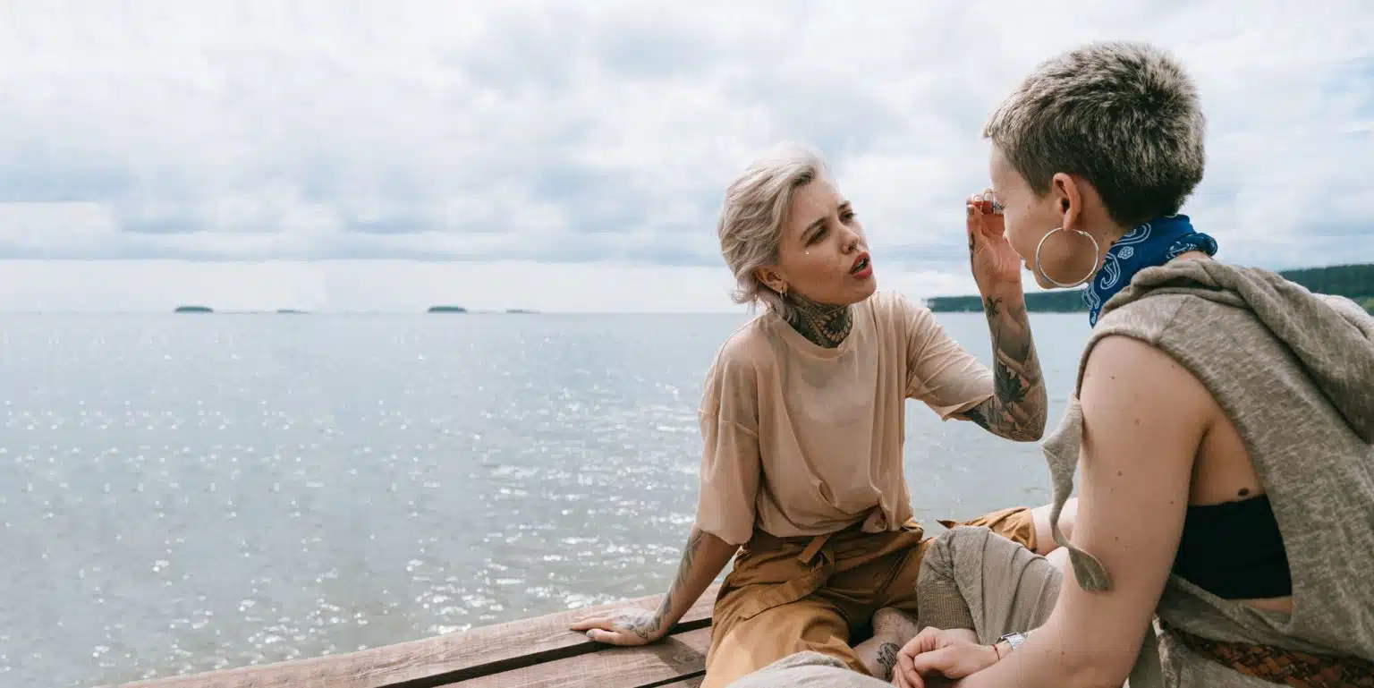 Two women with tattoos sitting on a pier one with her hand reaching for the face of the other