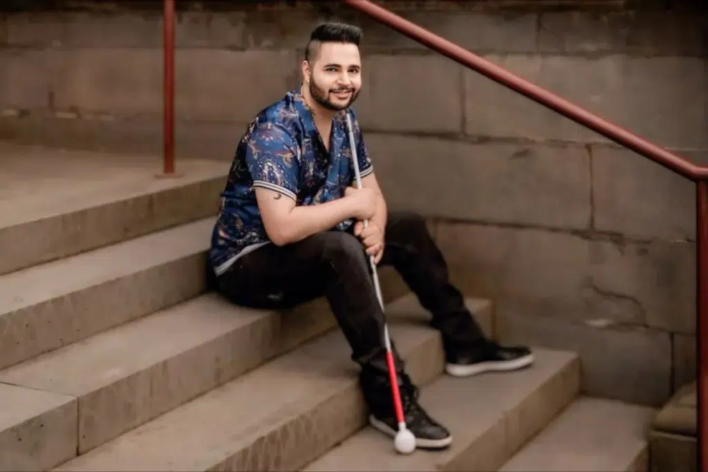 Karan Nagrani sitting on a stair with his cane, wearing a blue shirt and black jeans.