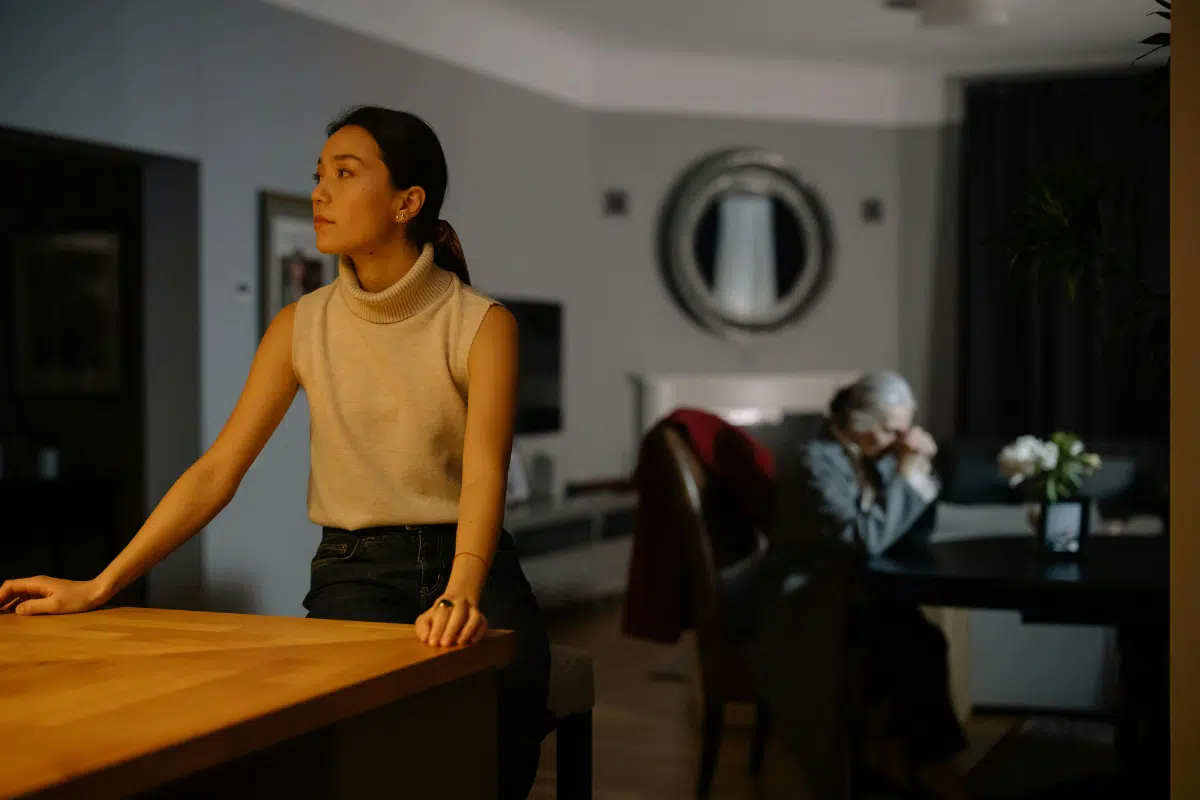 older woman standing at kitchen bench, mother looking sad at table in background